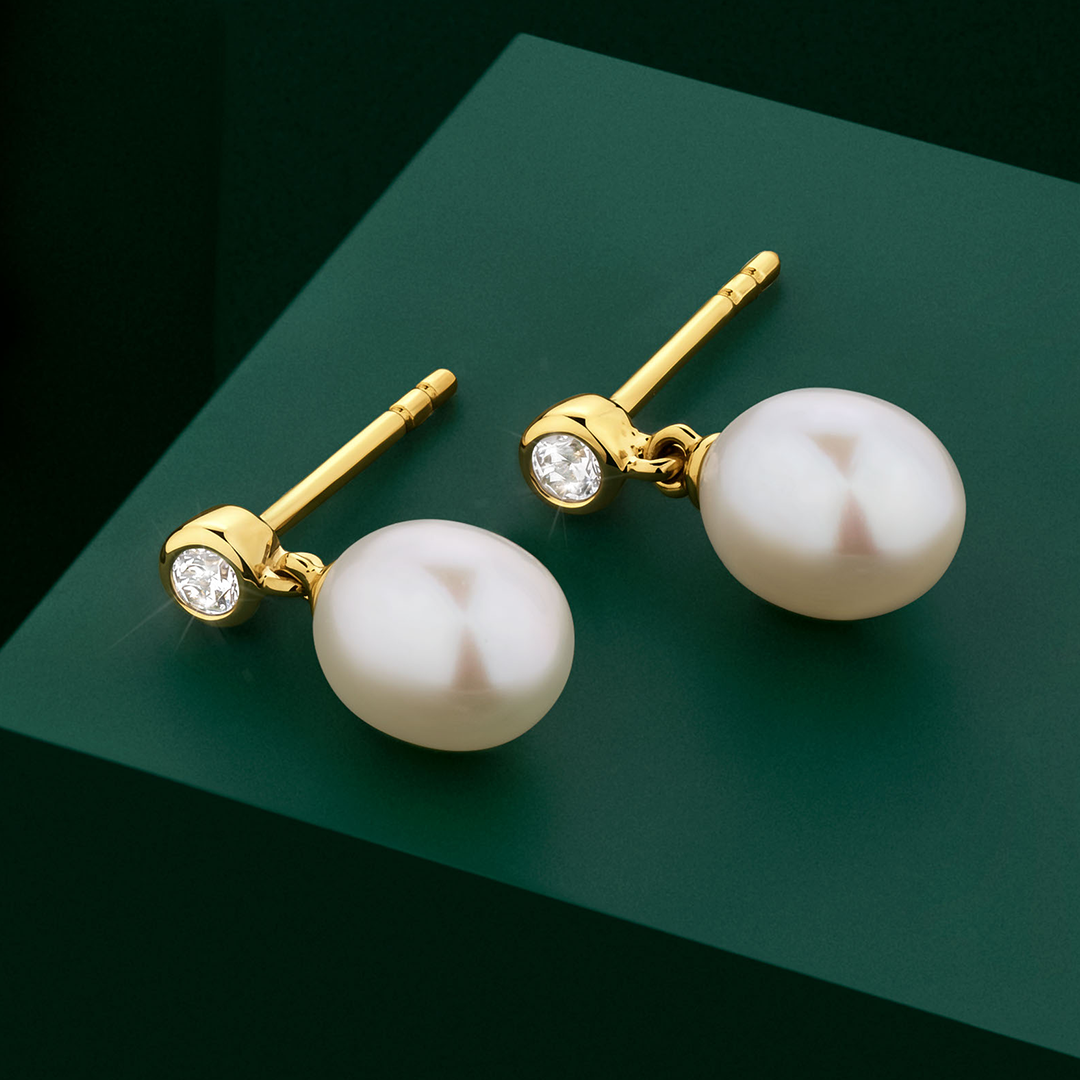 The Wallace Bishop Freshwater Pearl & Cubic Zirconia Bezel Drop Earrings in 9ct Yellow Gold, reference 33ECZ06Y, displayed on a pedestal. Each earring features a bezel-set round cubic zirconia stud with an 8x6mm freshwater pearl drop, 12.40mm in length, with butterfly backs.