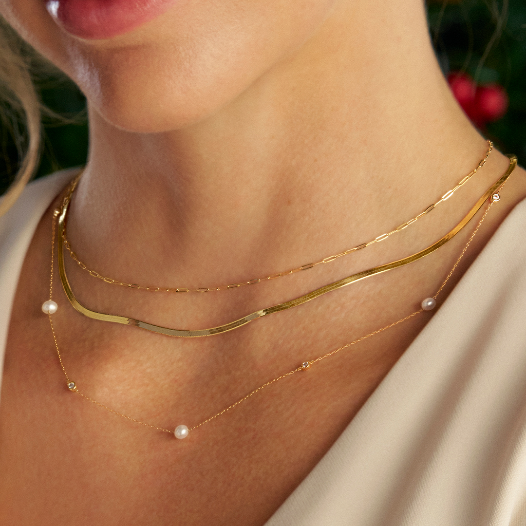 A neckline wearing two Wallace Bishop necklaces in 9ct Yellow Gold layered together: the Freshwater Pearl & Cubic Zirconia Bezel Necklace, reference 33PDCZ06Y, 50cm (19.7 inch) in length, with four round white freshwater pearls and four round cubic zirconias set at intervals along a cable style link chain, and the Two-Layer Necklace, reference 46KA2582, 500mm (19.7 inch) in length, pairing a flat herringbone chain with a paperclip link chain.