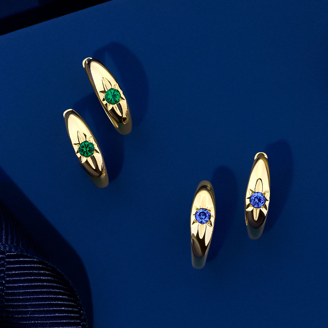 Front-on view of two pairs of Wallace Bishop Starburst Huggies in 9ct Yellow Gold, each measuring 14.50mm in length: the Created Emerald Starburst Huggies, reference 34ERSBEY, and the Created Sapphire Starburst Huggies, reference 34ERSBSY. Each huggie holds a 2.5mm round created gemstone, green emerald or blue sapphire, in a star hammer setting.