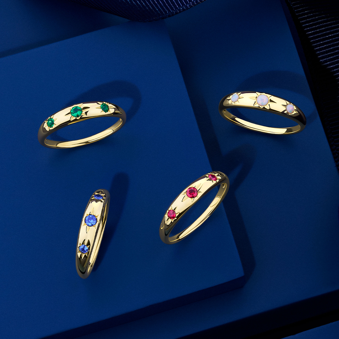 Front-on view of four Wallace Bishop Starburst Rings in 9ct Yellow Gold, each with three round gemstones in star hammer settings across a tapered band that narrows from 4mm to 2mm: the Opal Starburst Ring, reference 36RGSBOYO, the Created Emerald Starburst Ring, reference 38RGSBEYO, the Created Ruby Starburst Ring, reference 38RGSBRYO, and the Created Sapphire Starburst Ring, reference 38RGSBSYO.