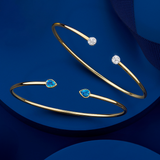 Front-on view of two Wallace Bishop flexi bangles in 9ct Yellow Gold on a plain background: the 0.15 TW Diamond Pavé Flexi Bangle, reference 3BG015Y, with pavé set diamonds, and the London Blue Topaz Flexi Bangle, reference 93BGLTBY, a cuff-style bangle measuring 50mm by 60mm with a bezel-set 5x4mm natural pear-cut London blue topaz on each side.