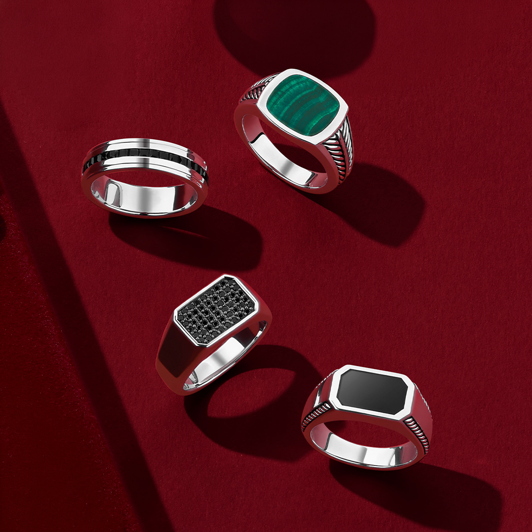 Flat lay view of four gents rings in sterling silver: the Hardy Brothers Princess-Cut Black Spinel Gents Ring, reference 4URG5BSSSV, 6.5mm in width, with a channel-set stripe of princess-cut black spinel through the middle, the Hardy Brothers Malachite Gents Signet Ring, reference 4URRP002V, with a cushion-shaped green malachite and rope detailing on the shoulders, the Wallace Bishop Elongated Onyx Gents Signet Ring, reference 4URG1BASSV, with a 15mm by 10mm octagonal black onyx and rope detailing on the sh