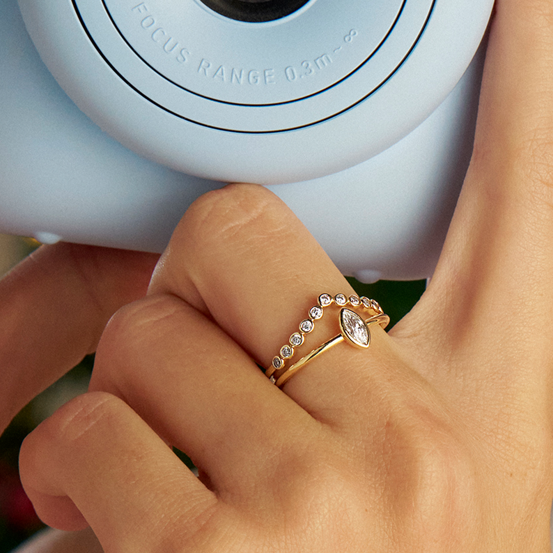 A finger wearing two Wallace Bishop cubic zirconia rings in gold: the Marquise Cubic Zirconia Ring, reference 68MQR01O, with a bezel-set 6x3mm marquise cut white cubic zirconia on a plain polished gold band, and the Cubic Zirconia Chevron Ring, reference 68RGCZY05N, in yellow gold with bezel-set round cubic zirconias set all along the front of the chevron.