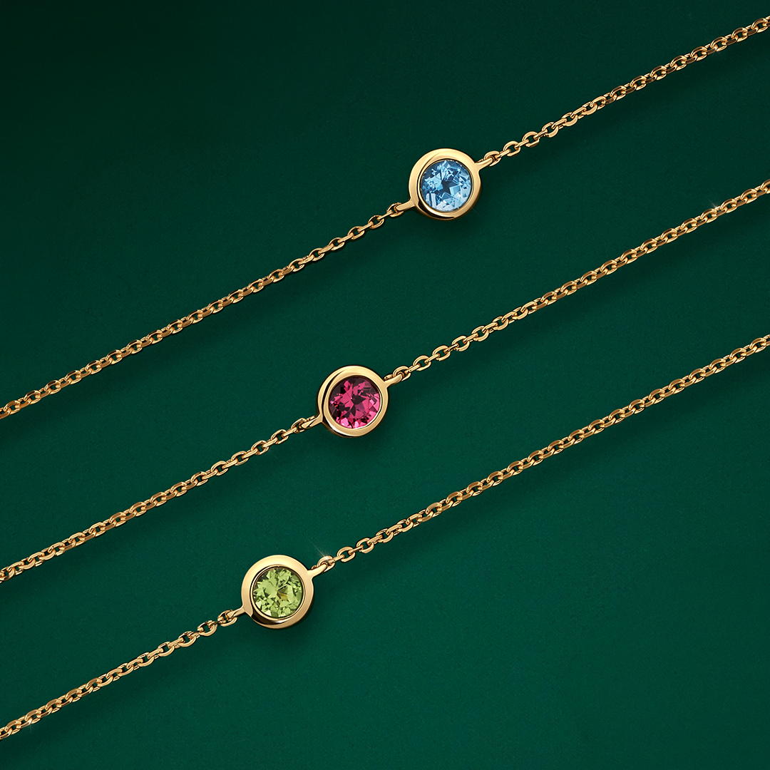 Flat lay close-up of the gemstone on three Wallace Bishop Bezel Bracelets in 9ct Yellow Gold, each on a fine cable chain: reference 93BLSBT3Y with a bezel-set 3mm round brilliant cut Swiss blue topaz, reference 93BLRDL3Y with a bezel-set 3mm round brilliant cut rhodolite garnet, and reference 93BLPER3Y with a bezel-set 3mm round brilliant cut peridot.