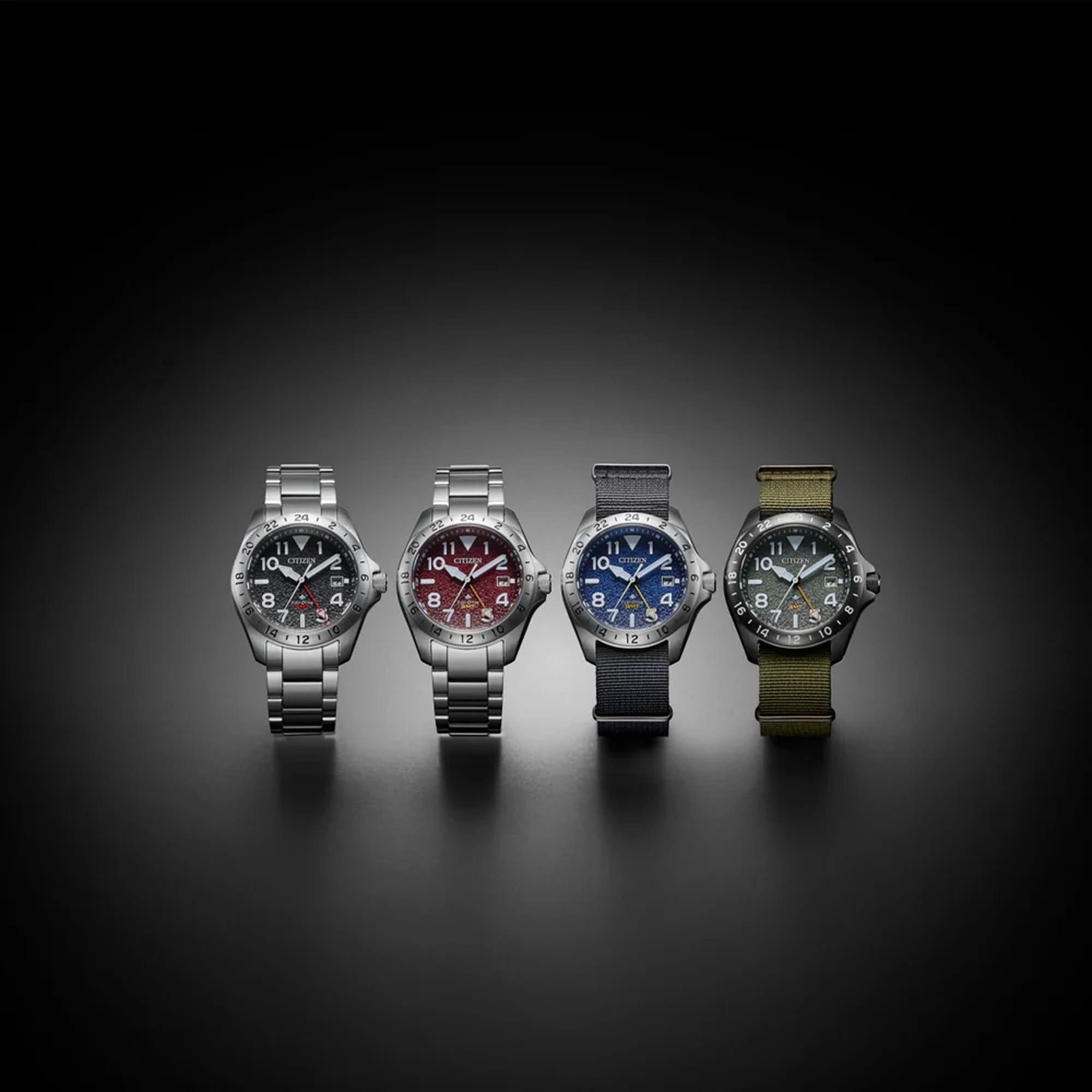 Four wristwatches with silver-toned metal cases are lined up against a dark gradient background. From left to right, the dials feature black, red, blue, and olive green colorways with matching bezels and straps.