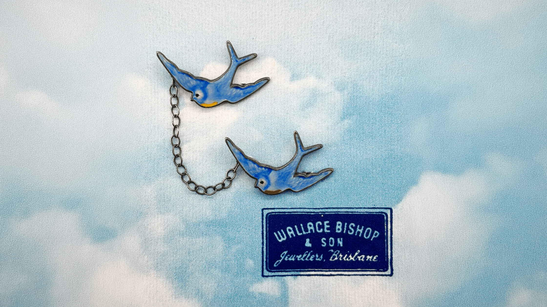 Two blue bird-shaped pins connected with a chain on a background with 'Wallace Bishop & Son' label.