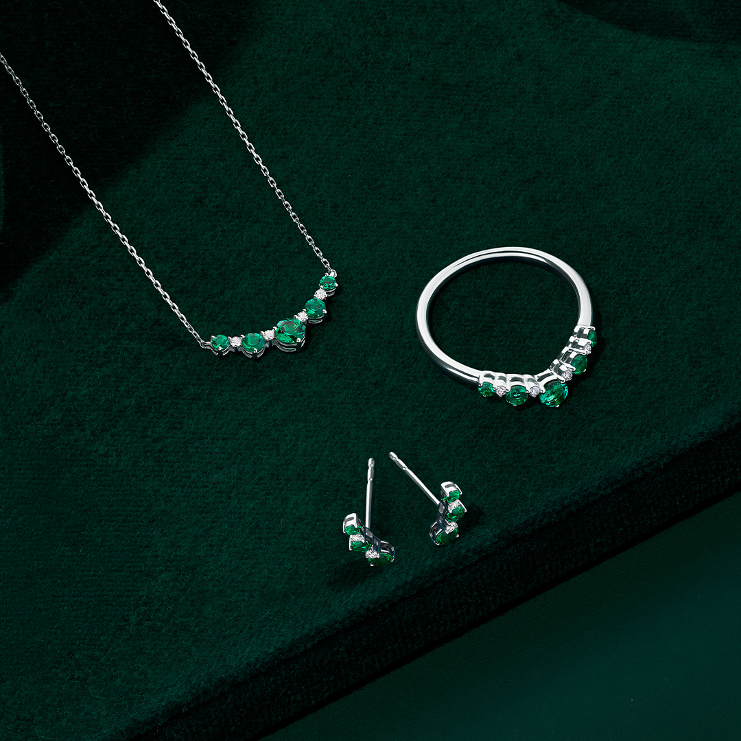 Front-on view of three Wallace Bishop Created Emerald & Lab-Grown Diamond Chevron pieces in Sterling Silver: the 0.04 TDW Pendant, reference LG33EME03, the 0.04 TDW Climber Earrings, reference LG33EME04, and the 0.04 TDW Ring, reference LG33EME02O. Each piece features round created emeralds with small round lab-grown diamonds set between them.