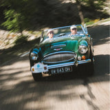 A vintage green convertible sports car driving on a sunlit, tree-lined road, with two people visible inside. The photo has a motion-blur effect due to speed, and the car front grille, headlights, and license plate are clearly visible.