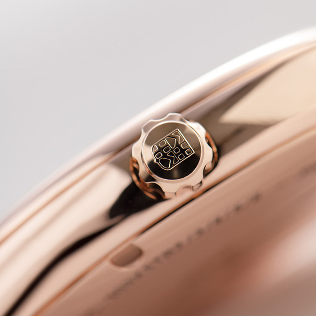 Close-up of a rose gold watch case showing the crown guard and part of the dial, with a soft gray background.