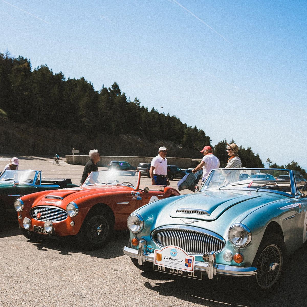 A group of classic convertible sports cars parked outdoors on a sunny day, with several people standing nearby. The most prominent car in the foreground is a light blue convertible, followed by a red convertible and other cars partially visible behind it.