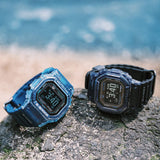 Two Casio G-LIDE GBXH5600-1D Black Digital Watches resting on a rocky surface in an outdoor environment. Rugged square case construction with black resin strap and multi-function digital dial on display.