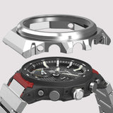 A close-up of a rugged analog watch with a black textured dial, silver-tone bezel, and red accents on the side, shown against a light gray background.