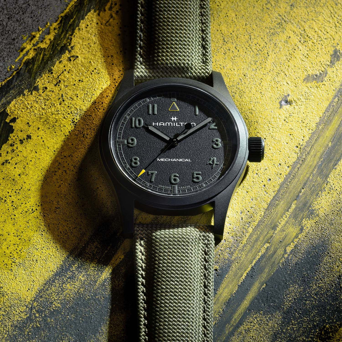 Hamilton x Call of Duty Khaki Field Mechanical Men’s 38mm Watch H69469930 is shown in close up, revealing the textured black dial, white Arabic numerals and subtle railroad minute track. The yellow triangle at 12 o’clock matches the yellow tip of the seconds hand.