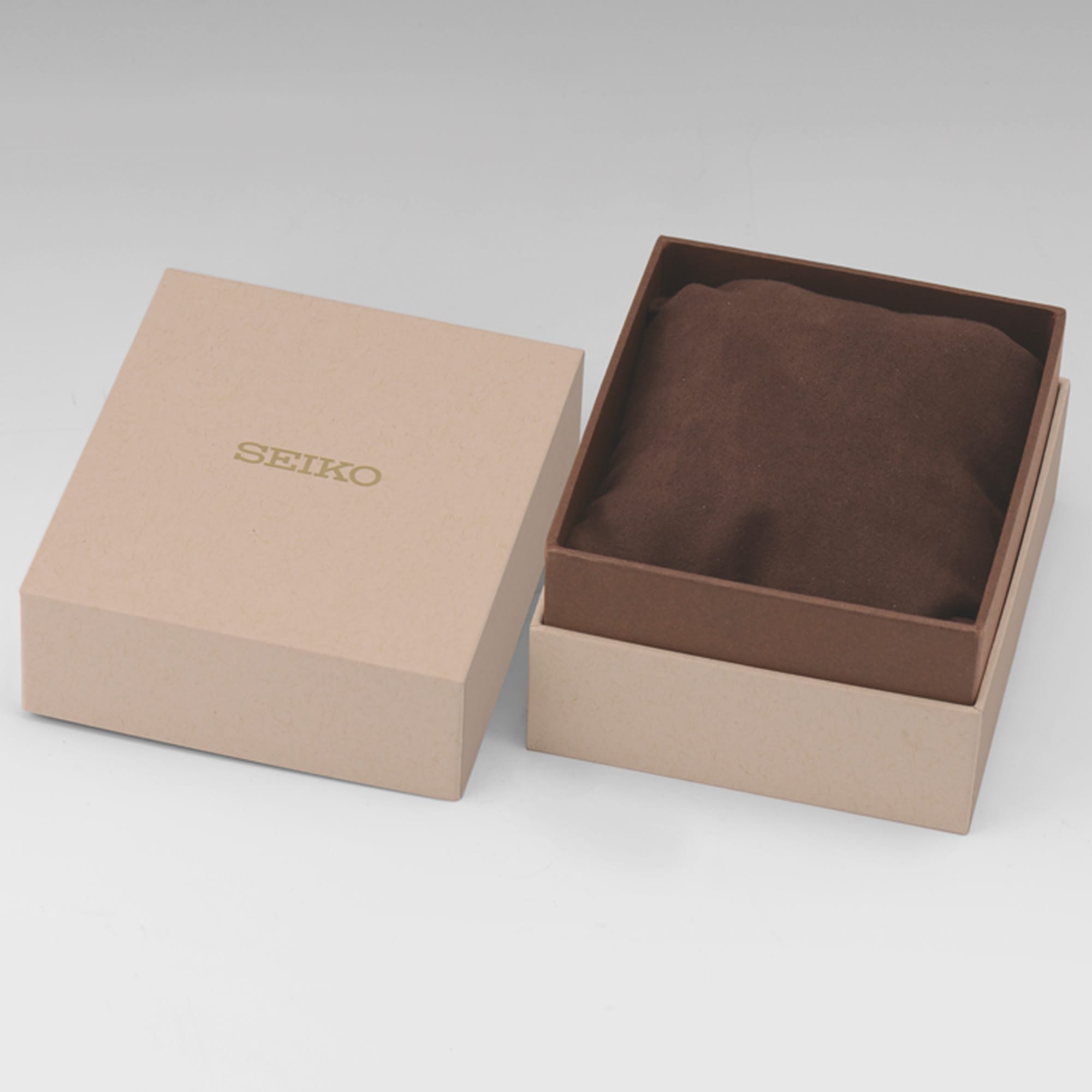 A Seiko branded presentation box with the lid off, showing a brown suede-like cushion inside. The box is a light tan/kraft color with gold 'SEIKO' lettering on the lid, photographed on a neutral light gray background.