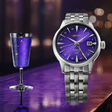 A Seiko Presage Cocktail Time Automatic GMT Men's 40.50mm Watch HCB006Jwith a purple sunburst dial and silver stainless steel bracelet, displayed next to a purple wine glass with gold leaf accents on a reflective purple surface.