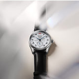 A Seiko automatic watch with a silver stainless steel case, white dial, black leather strap, and black Arabic numeral hour markers, shown against a soft gray background with light streaks.