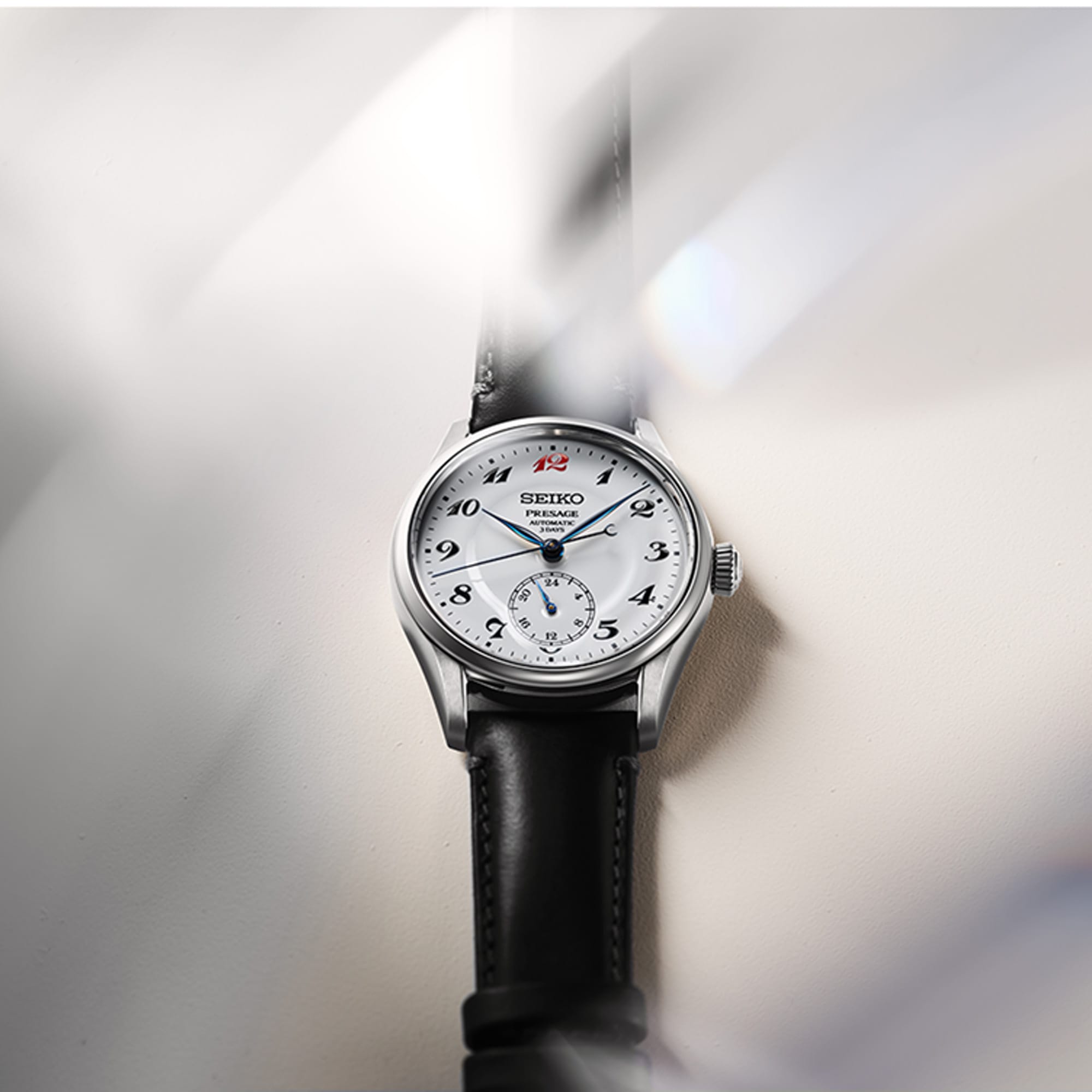 A Seiko automatic watch with a silver stainless steel case, white dial, black leather strap, and black Arabic numeral hour markers, shown against a soft gray background with light streaks.