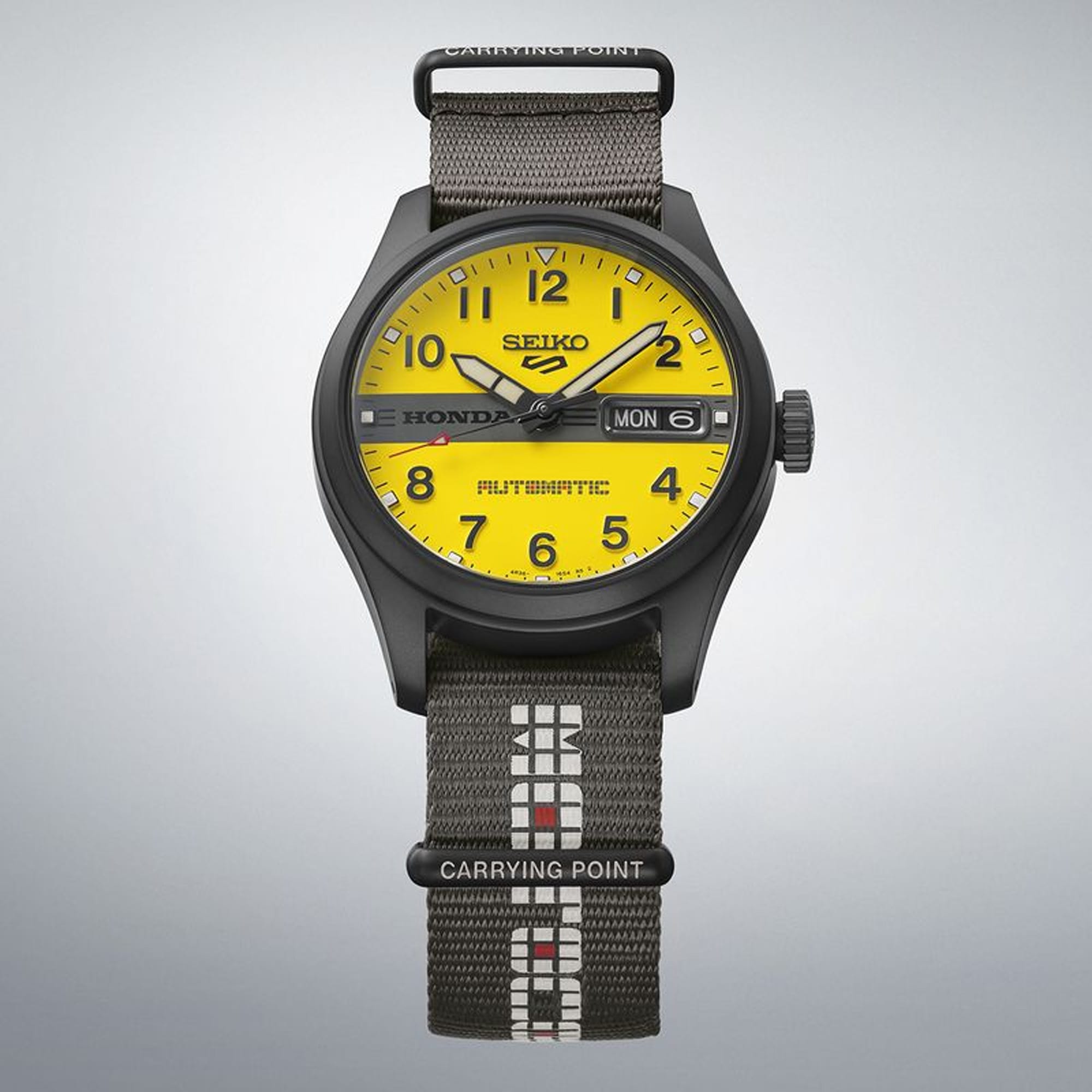 A Seiko Honda Automatic watch with a bright yellow dial, black case, and a dark gray woven fabric strap with white and red accents, shown against a light gray gradient background.