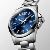 Three-quarter angle view of the Longines Conquest Men's 43mm Stainless Steel Quartz Watch L3.760.4.96.6, showing the case profile and screw-in crown. The angle highlights the sunray blue dial catching the light alongside the stainless steel bracelet.
