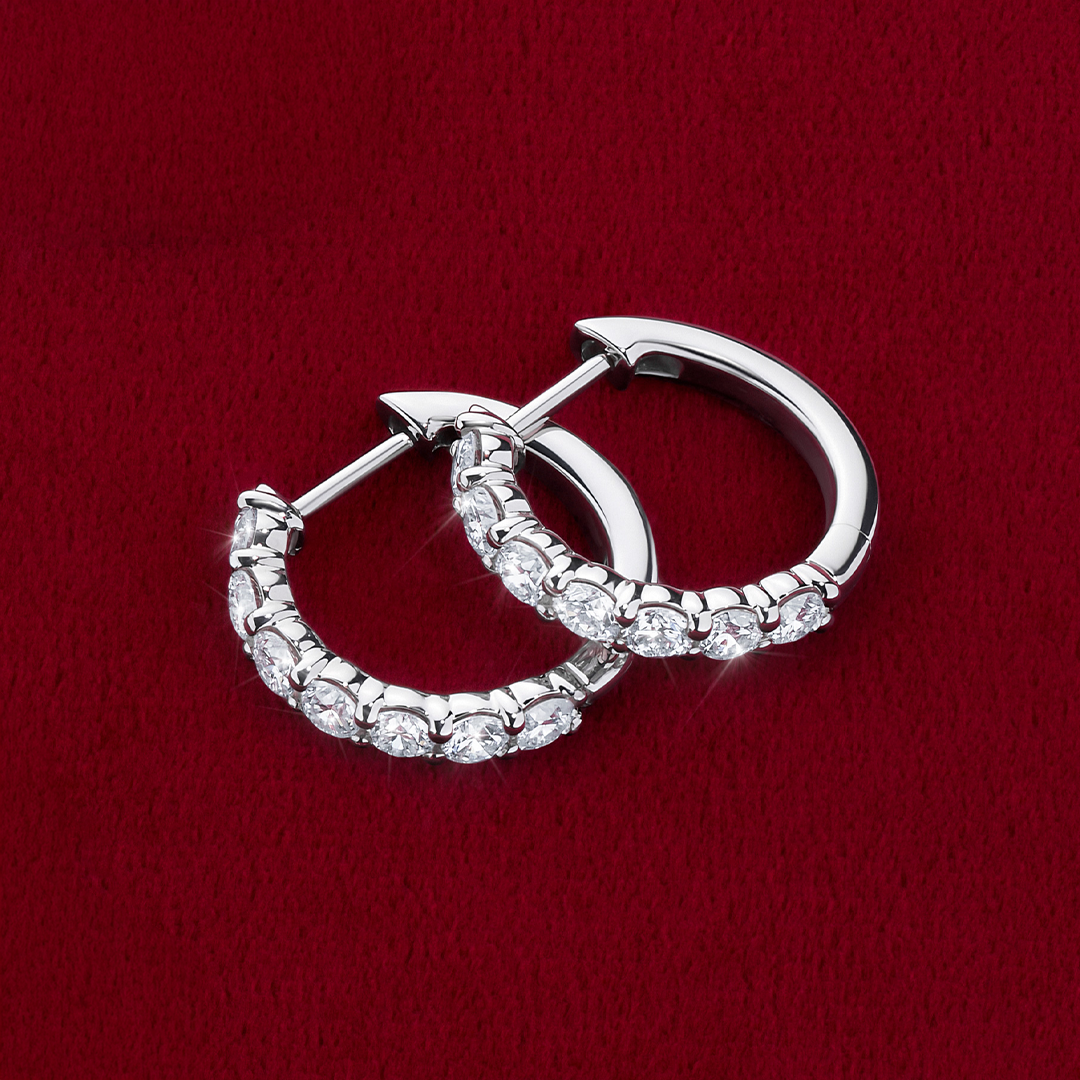 Flat lay view of the Wallace Bishop 1.00 TDW Round Lab-Grown Diamond Huggies in Sterling Silver, reference LGHUG04SS, laid on a deep red velvet surface. Each polished hoop is set with a row of round lab-grown diamonds along the front.