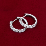 Flat lay view of the Wallace Bishop 1.00 TDW Round Lab-Grown Diamond Huggies in Sterling Silver, reference LGHUG04SS, laid on a deep red velvet surface. Each polished hoop is set with a row of round lab-grown diamonds along the front.