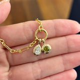 A hand holding two Wallace Bishop Double Bezel Pendants in 9ct Yellow Gold: the 0.30 TDW Pear-Cut Lab-Grown Diamond Double Bezel Pendant, reference LGNDBP30, and the Round Peridot Double Bezel Pendant, reference 93PDRDPE1Y, with the pear-cut diamond and the round green peridot each set in a polished gold double bezel.