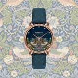 Morris & Co August Bera watch with a dark blue leather strap and a highly detailed, colorful peacock-themed dial, set against a William Morris-style floral and bird patterned background.