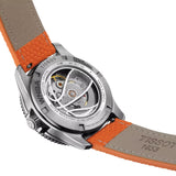 Close-up rear view of a Tissot watch with a silver-tone case, open back showing the mechanical movement, and an orange-and-tan leather strap on a white background.