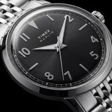 Close-up of a Timex Marlin wristwatch with a black dial, silver-tone stainless steel case, and matching metal link bracelet on a dark background.
