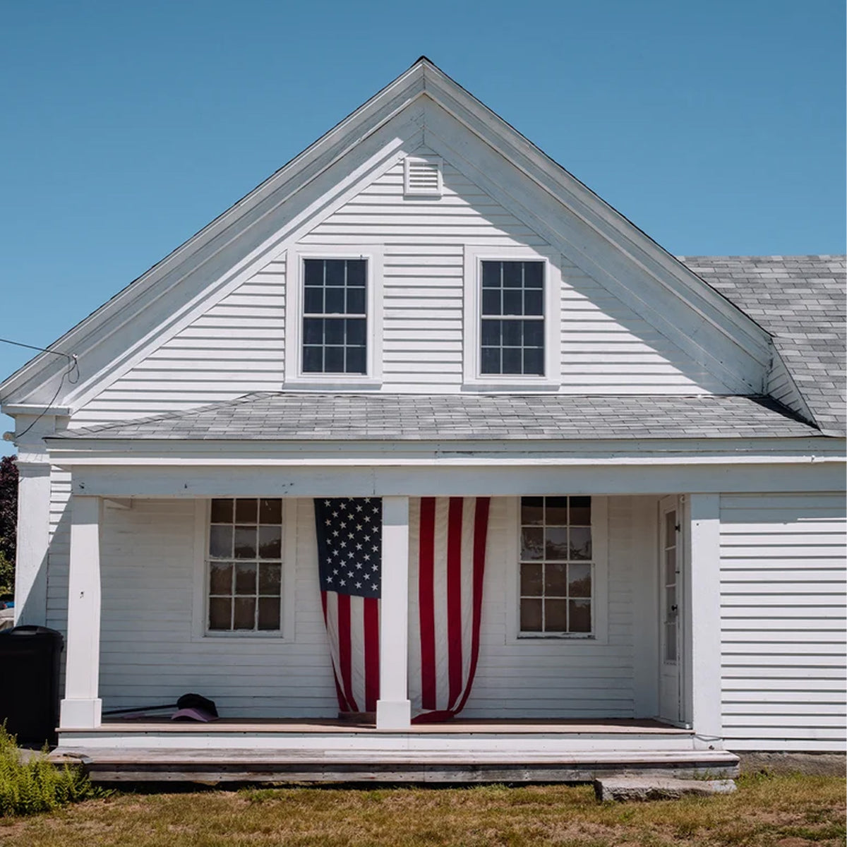 A white wooden house with a gray shingled roof, front porch columns, and an American flag hanging from the porch post. The house is photographed from the front under a clear blue sky.