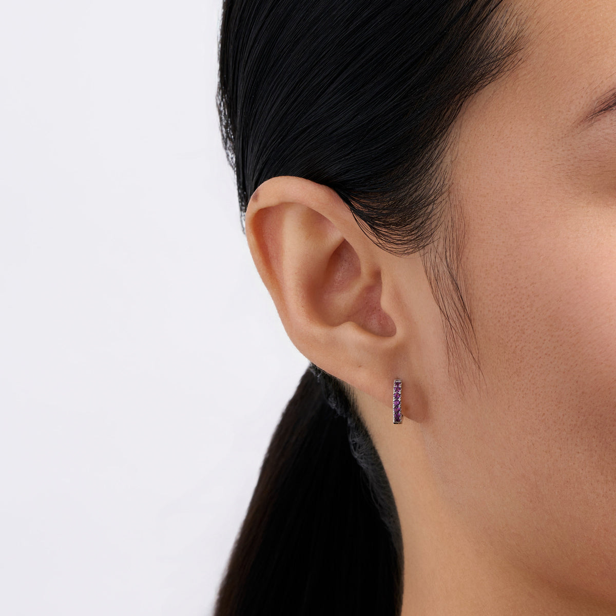 On-body view of a model wearing the Wallace Bishop Purple Cubic Zirconia Huggies in Sterling Silver, 10mm in size, hugging the earlobe with a line of purple cubic zirconias.