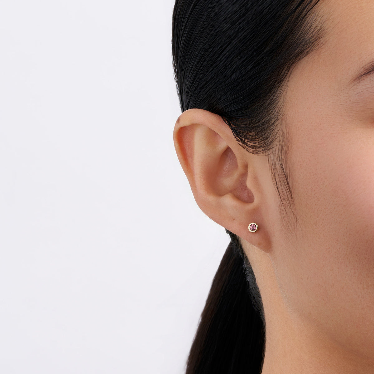 On-body view of a model wearing the Wallace Bishop Rhodolite Garnet Bezel Stud Earrings in 9ct Yellow Gold, with a 3mm round rhodolite garnet in each earlobe.