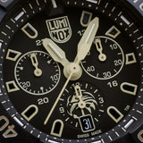 Close-up of the black dial on the Luminox Navy SEAL Foundation Men's 45mm Quartz Chronograph Watch XS.3590.NSF.SET, showing the sand-coloured details, three chronograph sub-dials and the Navy SEAL Foundation logo at 6 o'clock. This Navy SEAL Foundation Chronograph features luminous hands and markers with self-powered tritium tubes.