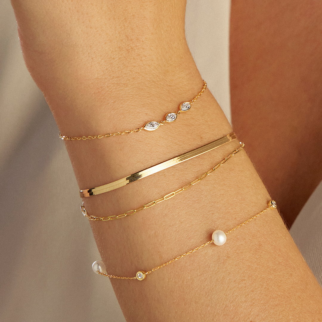 A wrist wearing a stack of three Wallace Bishop bracelets in 9ct Yellow Gold: the Freshwater Pearl & Cubic Zirconia Bezel Bracelet, reference 33BLCZ06Y, on a 190mm cable chain with stationed round white freshwater pearls and bezel-set cubic zirconias, the Marquise Cubic Zirconia Bezel Bracelet, reference 68MQB01, with three bezel-set marquise cubic zirconias on a 190mm fine chain, and the Two-Layer Bracelet, reference 45KA2582, with herringbone and paperclip link chains layered together, 210mm in length.