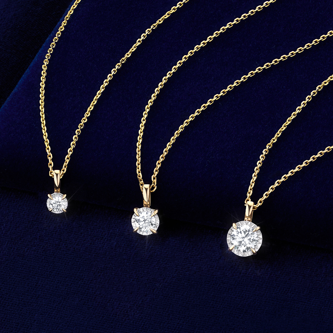 Flat lay view of three Wallace Bishop Essence Round Diamond Solitaire Pendants in 9ct Yellow Gold, each on a cable chain with a parrot clasp: the 0.07 TDW pendant, reference 3PBRI008Y, measuring 10mm in length, the 0.23 TDW pendant, reference 3PBRI023Y, and the 0.50 TDW pendant, reference 3PBRI05Y, measuring 11.30mm in length. Each pendant features a single natural round diamond in an illusion setting, hanging from a bale.