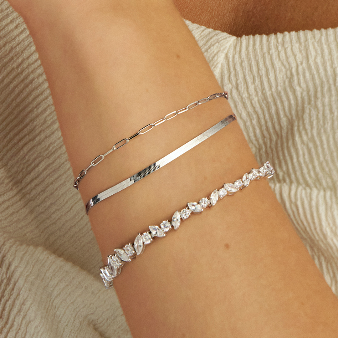 A wrist wearing a stack of two Wallace Bishop bracelets in Sterling Silver: the Cubic Zirconia Marquise & Round Cut Tennis Bracelet, reference 41BCZ908, with a line of marquise and round cut colourless cubic zirconias, and the Two-Layer Bracelet, reference 32KA2582, 210mm in length with an adjustable slider, pairing a herringbone chain with a paperclip link chain.