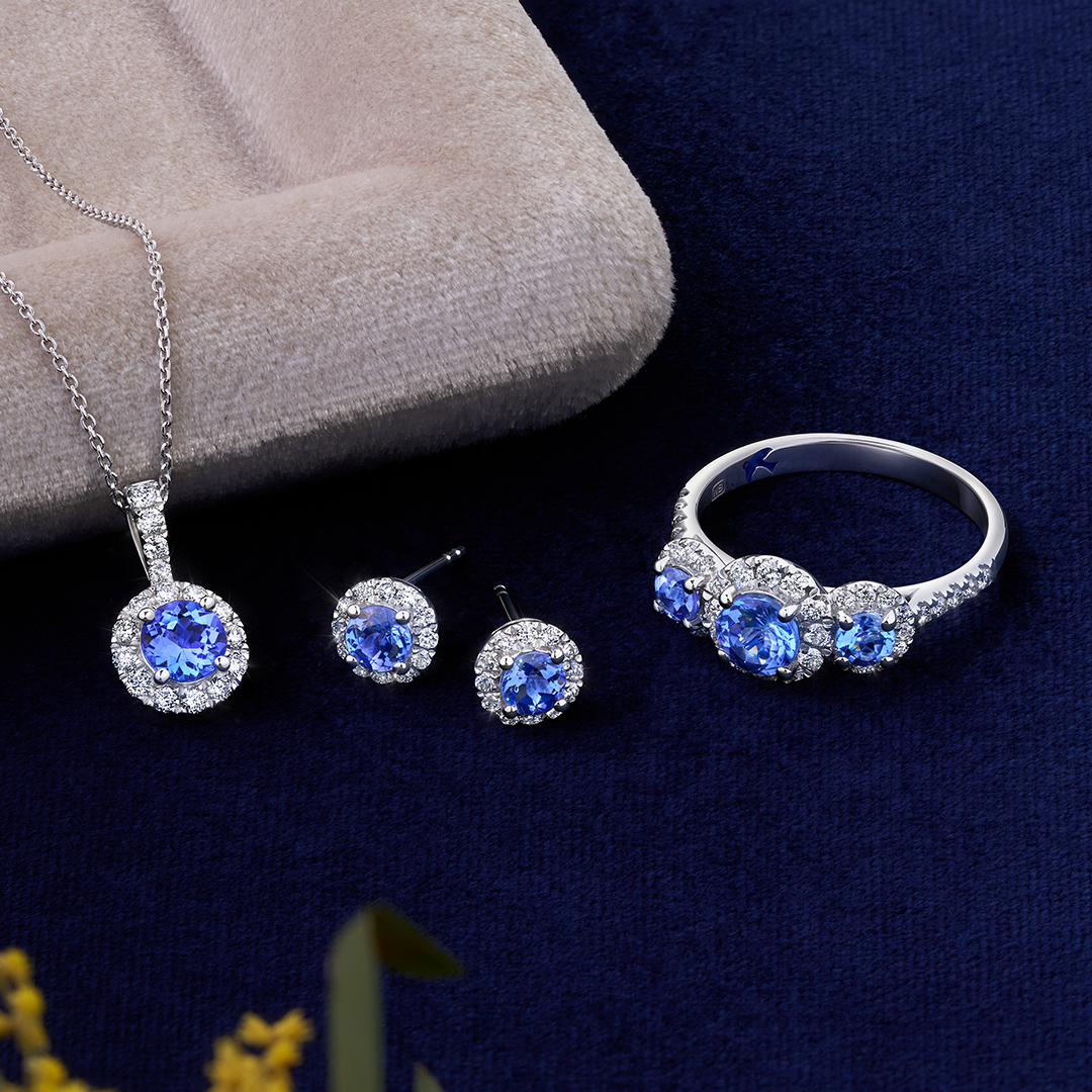 Front-on view of three Wallace Bishop Bluebird tanzanite pieces in 9ct White Gold: the 0.38 TDW Round Tanzanite & Diamond Halo Ring, reference 92RGTZ02WO, a trilogy ring with natural round tanzanites each surrounded by a halo of natural diamonds, with more diamonds on the shoulders, the 0.19 TDW Round Tanzanite & Diamond Halo Stud Earrings, reference 93ERTZ02W, each with a natural 4mm round tanzanite in a diamond halo, and the 0.23 TDW Round Tanzanite & Diamond Halo Pendant, reference 93PDTZ02W, 14.35mm in 