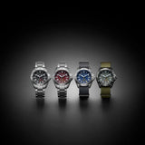 Four wristwatches with silver-toned metal cases are lined up against a dark gradient background. From left to right, the dials feature black, red, blue, and olive green colorways with matching bezels and straps.