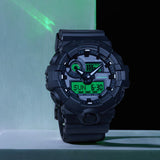 Casio G-Shock GA100BEG-1A front view showcasing the 55mm x 51.2mm black resin case with centre analogue display flanked by four large digital LCD windows with electro green printed accents, large aluminium hands, and the GA-100's signature oversized bold silhouette.