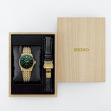 A Seiko watch presented in an open light wood box next to its lid. The watch has a gold-tone metal bracelet and case with a dark green dial and a black textured strap with a gold buckle.