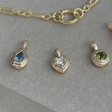 Flat lay view of three Wallace Bishop Double Bezel Pendants in 9ct Yellow Gold: the Pear-Cut Swiss Blue Topaz Pendant, reference 93PDPSST1Y, the 0.30 TDW Heart Lab-Grown Diamond Pendant, reference LGNDBH30, and the Round Peridot Pendant, reference 93PDRDPE1Y. Each pendant has a single gemstone set in a polished gold double bezel.