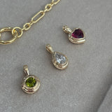 Flat lay view of three Wallace Bishop Double Bezel Pendants in 9ct Yellow Gold: the Heart Rhodolite Garnet Pendant, reference 93PDHTRH1Y, the Round Peridot Pendant, reference 93PDRDPE1Y, and the 0.30 TDW Pear-Cut Lab-Grown Diamond Pendant, reference LGNDBP30. Each pendant has a single gemstone set in a polished gold double bezel.