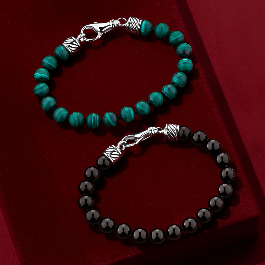 Two Wallace Bishop gents beaded bracelets in Sterling Silver laid on a deep red surface: the Malachite Gents Bracelet, reference 4UBLBASS2, with round green malachite beads, and the Onyx Gents Bracelet, reference 4UBLBASS3, with round black onyx beads. Each bracelet has sterling silver end caps with ridged detailing and a lobster clasp.