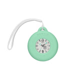 Casio Baby-G+PLUS BGA15K-7A front view — white matte round resin case and band, shimmering metal dial with green accents, Arabic numerals, three-hand analogue display with rotating day design. Includes green silicone bag charm holder. 43.5mm x 42.4mm, 35 grams, shock resistant, 100M water resistant.