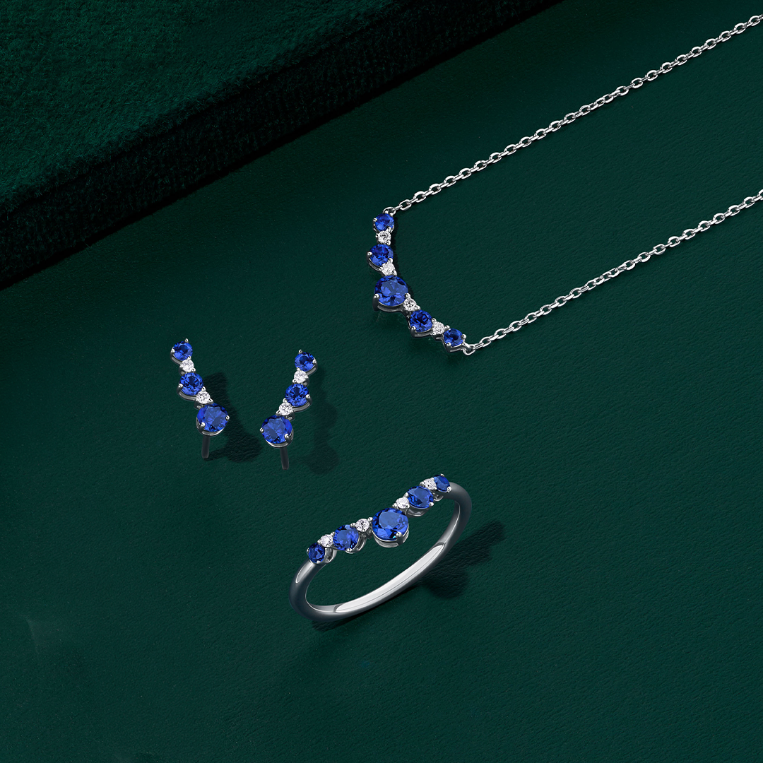 Front-on view of three Wallace Bishop Created Sapphire & Lab-Grown Diamond Chevron pieces in Sterling Silver: the 0.04 TDW Pendant, reference LG34SAP03, the 0.04 TDW Climber Earrings, reference LG34SAP04, and the 0.04 TDW Ring, reference LG38SAP02O. Each piece features round dark blue created sapphires with small round lab-grown diamonds set between them.