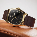 The Timex Marlin Chatham Automatic 36mm Watch TW2Y84500 is shown resting on a tan leather chair. The black dial, gold-tone case and brown leather strap bring a mid-century look to relaxed everyday outfits.