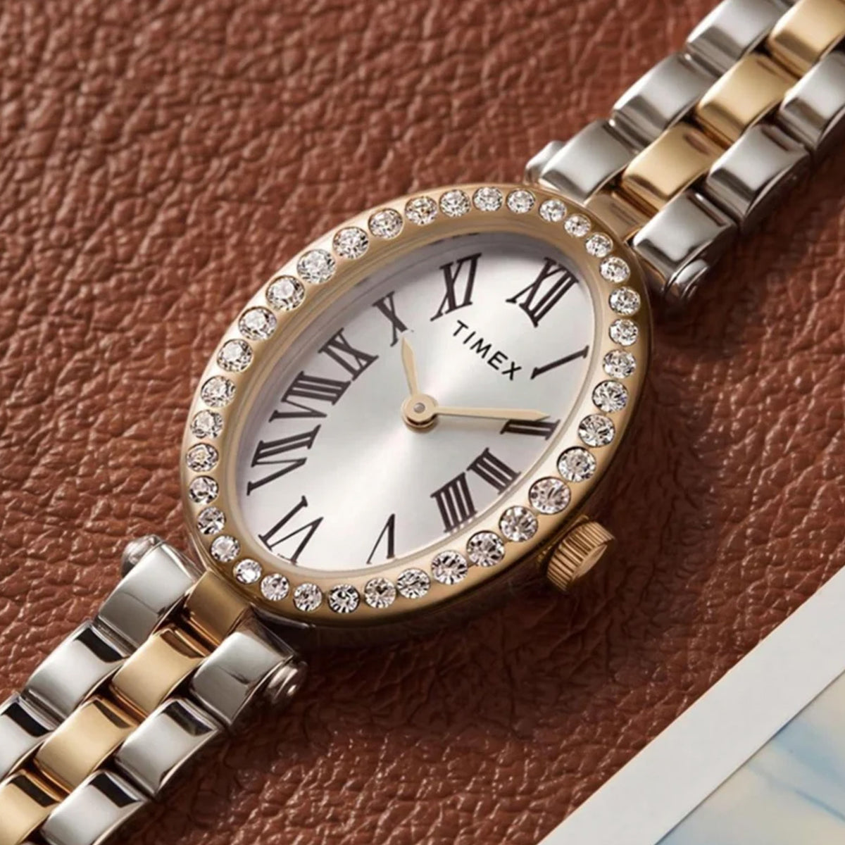 The Timex Cavatina Quartz 19mm Women’s Watch TW2Y90200 is shown resting on brown leather beside a photograph. The two-tone bracelet and crystal bezel give it a jewellery-like look.