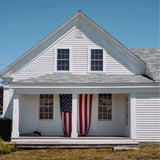 A white wooden house with a gray shingled roof, front porch columns, and an American flag hanging from the porch post. The house is photographed from the front under a clear blue sky.